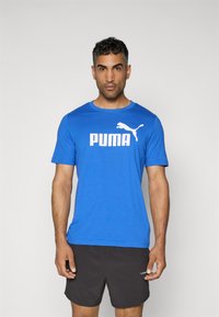 Zils īsām piedurknēm T-krekls ar baltu "PUMA" logo un kaķa emblēmu; samtaina auduma, apaļa kakla izgriezums, saskaņots ar melniem šortiem.