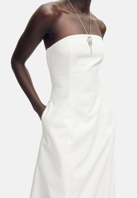 Robe blanche sans bretelles avec un corsage ajusté et des poches. Présente des lignes épurées et une texture lisse, agrémentée d un simple collier en argent.