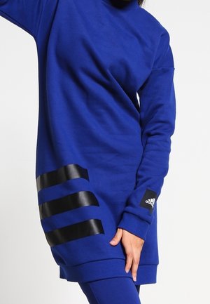 Osoba ubrana w długą niebieską bluzę Adidas z trzema czarnymi paskami przy krawędzi i logo Adidas na rękawie.