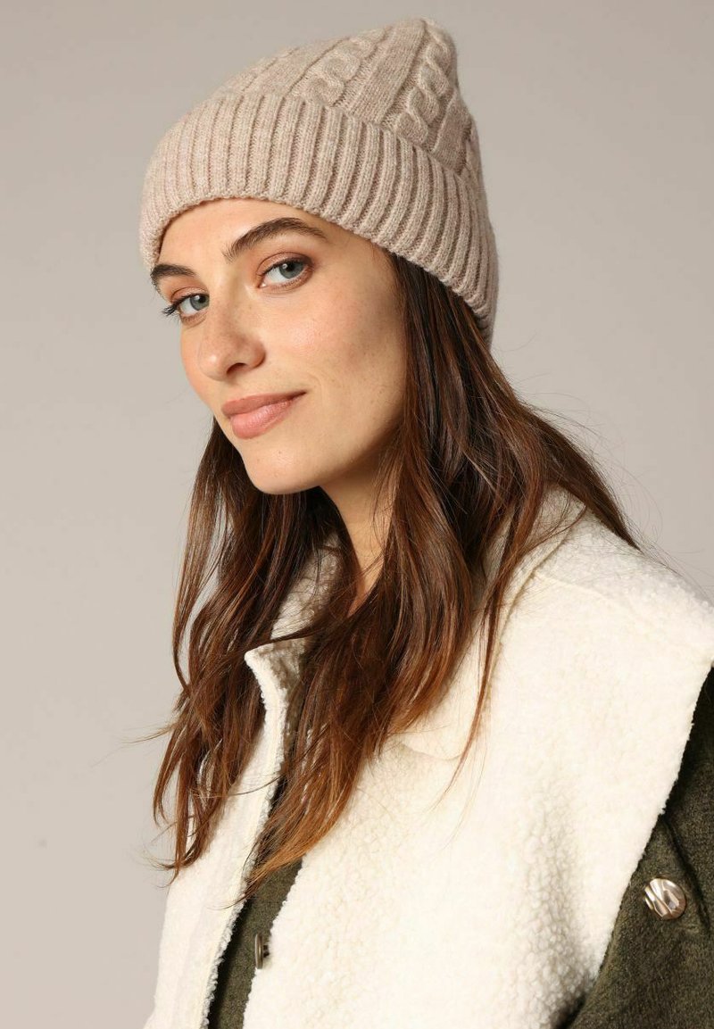 Bonnet tricoté en beige clair, avec un design en côtes et des motifs en câble. Texture douce et forme ajustée.