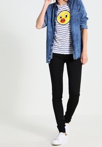 Camisa de mezclilla sobre una camiseta de rayas con un gráfico de emoji amarillo, combinada con jeans negros y zapatillas blancas. Atuendo casual.