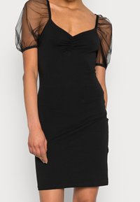 Robe noire à silhouette ajustée, dotée de manches bouffantes transparentes et d'un corsage froncé. Le tissu semble lisse avec une légère élasticité.