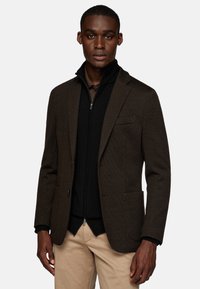 Brown textured blazer with a single chest pocket, paired with a black zip-up inner jacket, worn over a light shirt and beige pants.