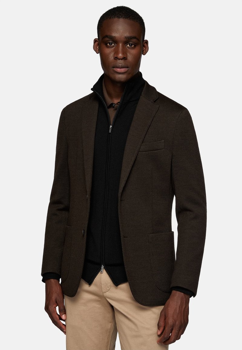 Brown textured blazer with a single chest pocket, paired with a black zip-up inner jacket, worn over a light shirt and beige pants.