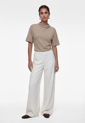 Donna in piedi che indossa una maglietta beige a maniche corte, pantaloni bianchi a gamba ampia, scarpe marroni e orecchini a cerchio, su uno sfondo chiaro semplice.
