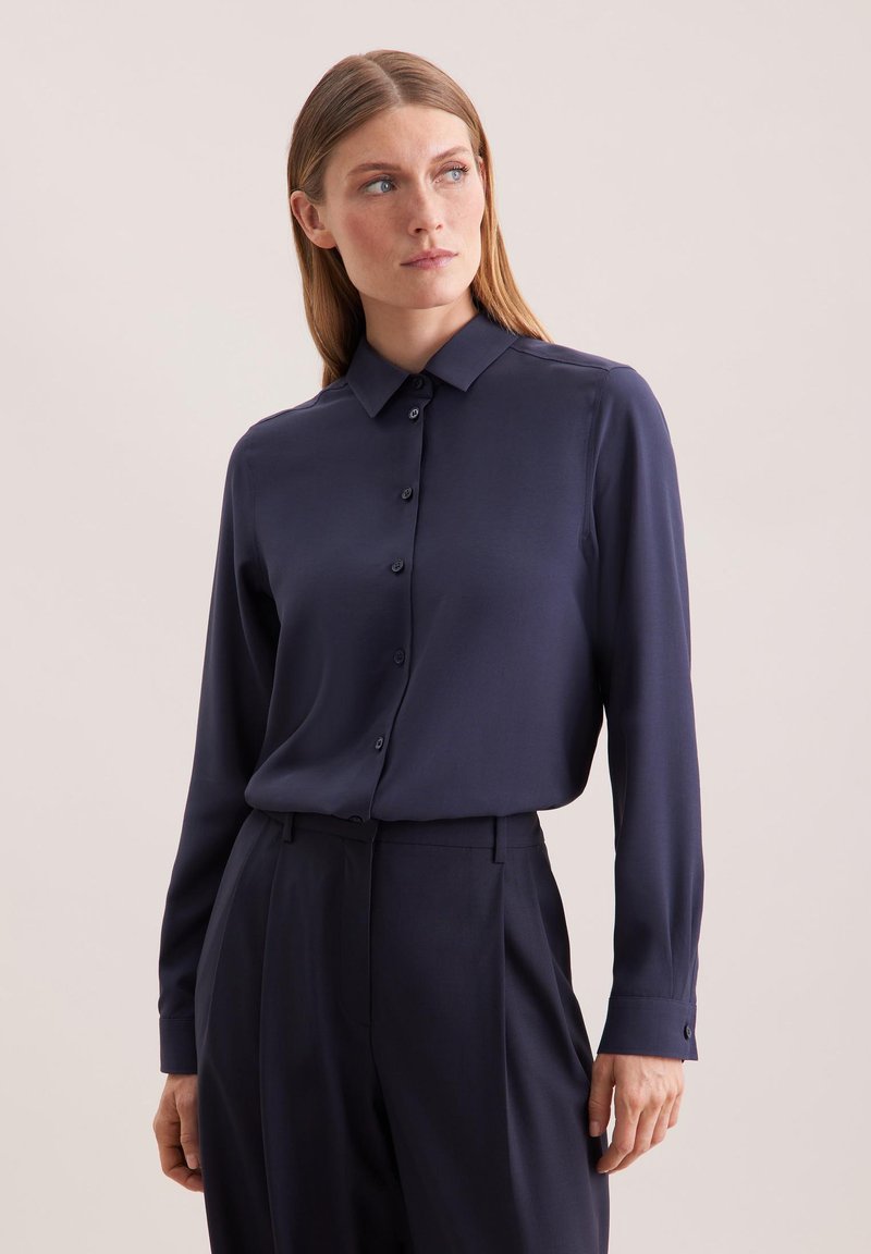 Femme aux cheveux châtain clair et raides, portant une chemise bleu marine foncé à manches longues boutonnée et un pantalon assorti, regardant sur le côté.