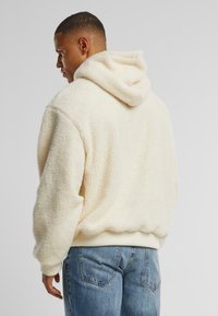Cremefarbener Kapuzen-Fleece-Pullover mit einer weichen, strukturierten Oberfläche, gerippten Bündchen und einer lässigen Passform. Kombiniert mit hellblauen Jeans.