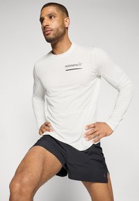 Hvit langermet treningsskjorte med logo på venstre bryst, kombinert med svarte shorts. Glatt, lett stoff, slim fit design.