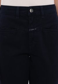 Taille de jean noir avec un bouton argenté et une étiquette de marque visible sur la zone de la fermeture éclair, portée avec un haut blanc.