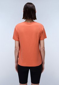 Kurze Ärmel, orange T-Shirt aus Baumwolle mit rundem Ausschnitt, lockerer Passform und einem kleinen Etikett auf der linken Seite. Kombinieren Sie es mit schwarzen Shorts.
