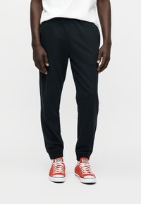 Μαύρο βαμβακερό παντελόνι jogging με ελαστική μέση, τελειώματα με μανσέτες, σε συνδυασμό με κόκκινα πάνινα αθλητικά παπούτσια με λευκές σόλες και κορδόνια.