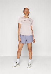 Camiseta desportiva rosa claro com logótipo, combinada com calções lilás. Meias brancas até ao tornozelo e sapatilhas cinzentas completam o visual.