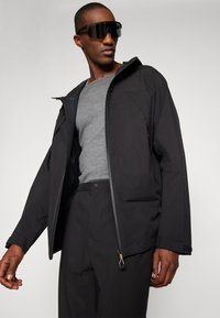 Veste imperméable noire avec fermeture éclair et ourlet ajustable, portée par-dessus une chemise grise. Le mannequin porte des lunettes de soleil noires surdimensionnées.
