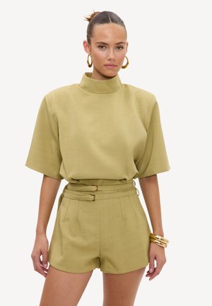 Femme avec les cheveux en chignon, portant un haut à manches courtes à col haut vert olive et un short taille haute assorti avec double ceinture et boucles d'oreilles créoles dorées.