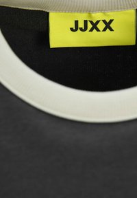 Černý tričko s límcem ve světle béžovém žebrovaném provedení. Jasně žlutá visačka s logem "JJXX" je viditelná. Hladká textura a minimalistický design.