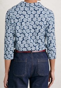 Granatowa bluzka w kwiatowy wzór z rękawami trzyczwartowymi; niebieskie kwiaty z białymi środkami; w zestawie z ciemnoniebieskimi dżinsami z dwoma tylnymi kieszeniami.