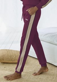 Bordeauxfarbene Sweatpants mit beigen Seitenstreifen und schwarzem Piping. Hergestellt aus weichem Stoff, mit elastischem Bund und Kordelzug.