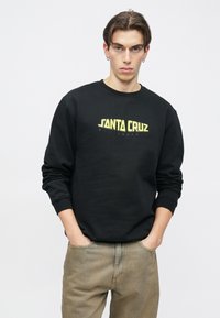 Černá mikina s žlutým logem "SANTA CRUZ" a textem "SKATEBOARD". Měkká látka, kulatý výstřih a žebrované manžety. Nošena s lehkými khaki kalhotami.