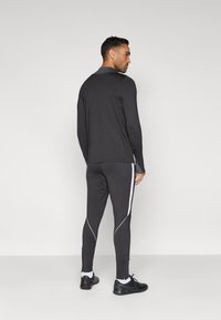 Μαύρο αθλητικό τοπ με μακριά μανίκια, συνδυασμένο με εφαρμοστά μαύρα παντελόνια με λευκές ρίγες στα πλάγια. Το υλικό φαίνεται λείο και με κομψό σχέδιο.