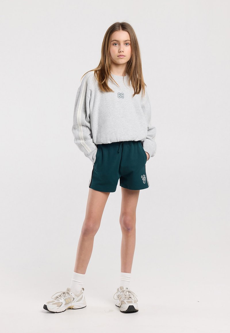 Joven niña de pie con las manos en los bolsillos, vistiendo una sudadera gris, pantalones cortos verde oscuro con logo, calcetas blancas y zapatillas blancas sobre fondo blanco.