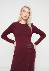 Bordo haljina s dugim rukavima, rebrastom teksturom, ukrasnim zakovicama oko vrata i dvostrukim kopčama u struku s detaljima na dugmadi.