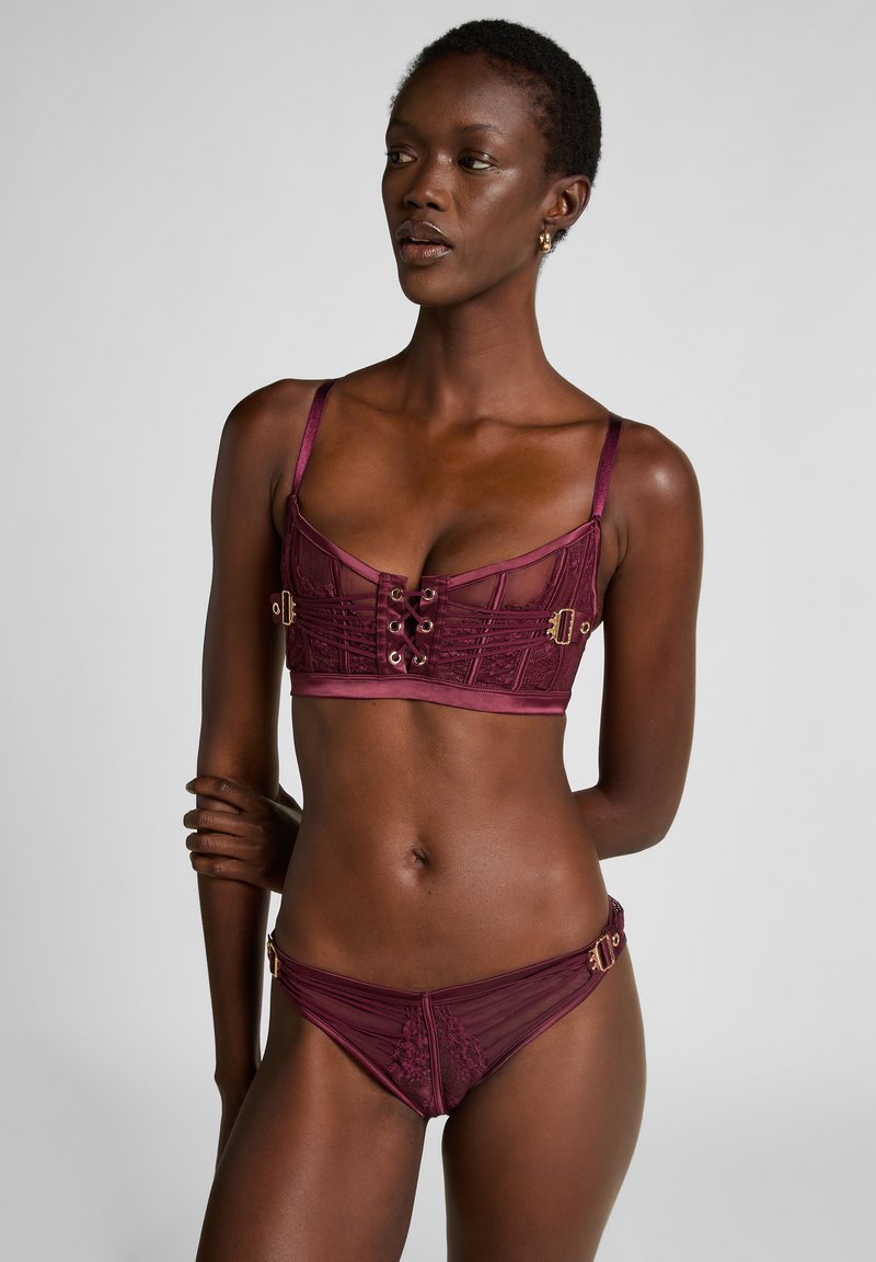 Donkere vrouw in bordeauxrode kanten lingerie met verstelbare bandjes, staand met gekruiste armen, kijkend opzij tegen een grijze achtergrond.