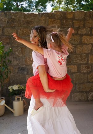 Zwei Personen in einer Gartenumgebung; eine trägt einen weißen gestuften Rock, die andere ein rosa Hemd und einen roten Tüllrock, beide lächeln.