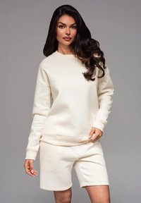Completo felpa e pantaloni corti color crema, realizzato in tessuto morbido, con polsini a coste, vestibilità rilassata e design minimalista senza motivi visibili.
