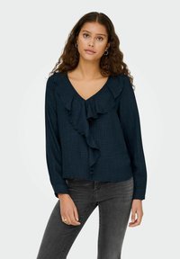 Navy blauwe blouse met lange mouwen, een v-hals en een gerimpelde voorkant. Gestructureerde stof met een subtiele glans. Draag met donkergrijze jeans.