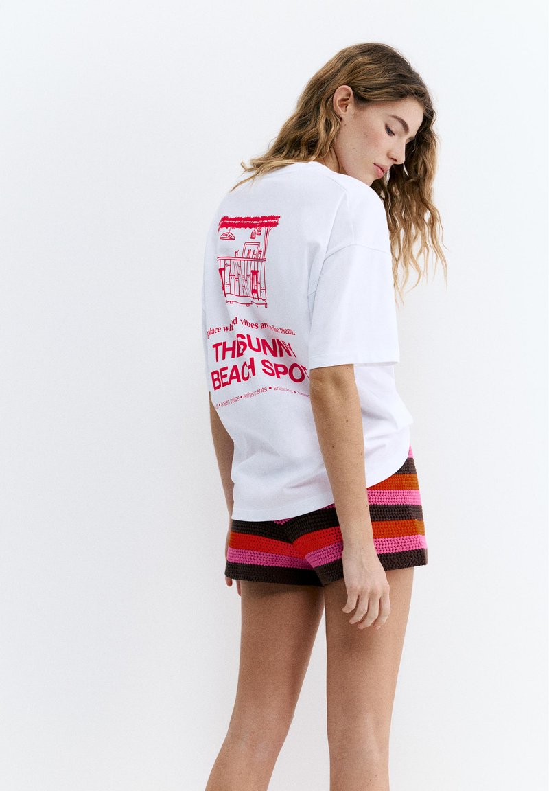 Jeune femme portant un T-shirt blanc oversize avec un imprimé rouge de bar de plage et un short en crochet rayé multicolore, debout devant un fond blanc.