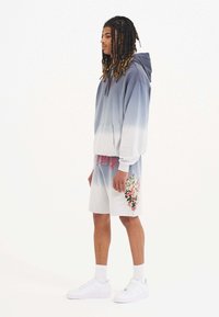 Sudadera con capucha en un degradado de azul a blanco, combinada con pantalones cortos blancos que presentan un gráfico colorido de un dragón. Zapatillas blancas completan el look.