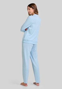 Mujer de pie descalza, mirando hacia la izquierda, vestida con una blusa de mangas largas de lunares azul claro y pantalones a juego, contra un fondo gris liso.