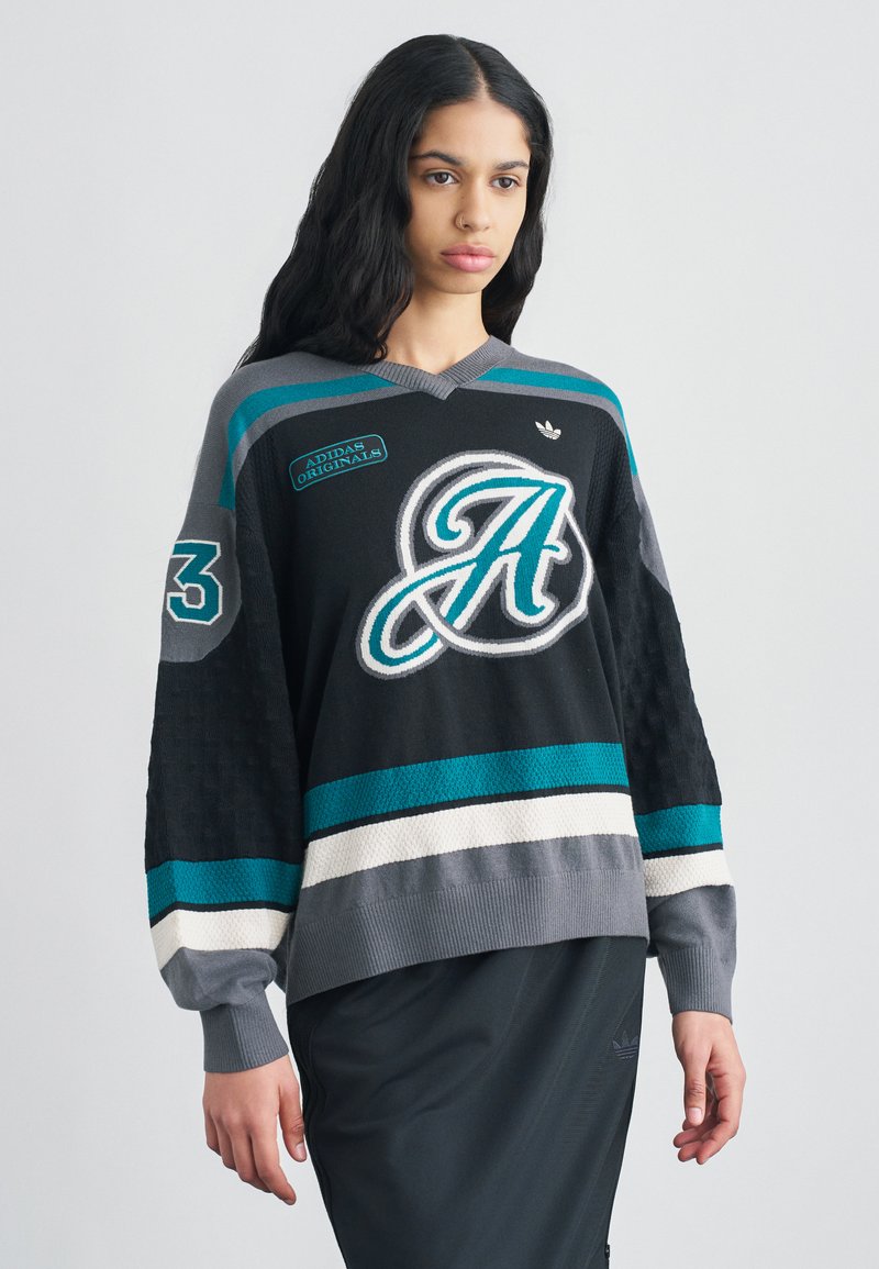 Maillot surdimensionné en maille noir et sarcelle avec un col en V, arborant la lettre "A" et des logos de la marque. Poignets côtelés et manches texturées.