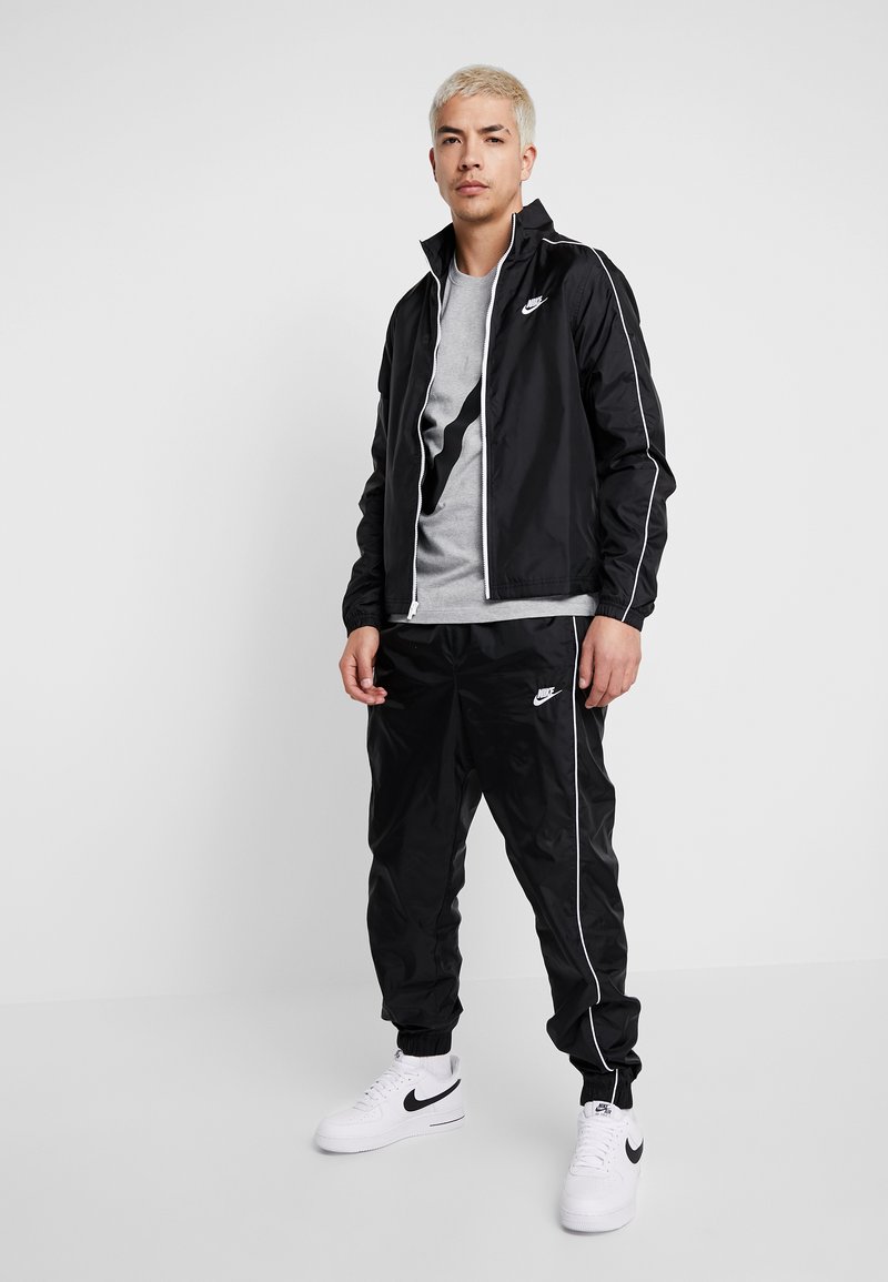 Melna Nike vēja jaka ar baltām akcentiem, kombinēta ar melniem treniņbikšu un pelēku T-kreklu. Iespējas ar rāvējslēdzēju un logotipa detalizāciju.