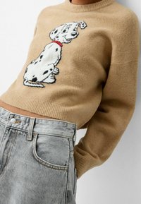 Beige gestrickter Pullover mit cropped Design, der ein schwarz-weißes Dalmatiner-Motiv und einen roten Kragen hat. Kombiniert mit grauen Jeansshorts.