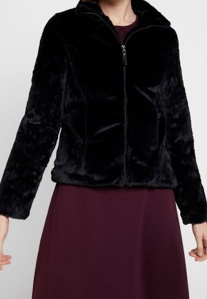 Personne portant une veste noire en peluche zippée sur une robe grenat, visible du cou aux hanches sur un fond uni.
