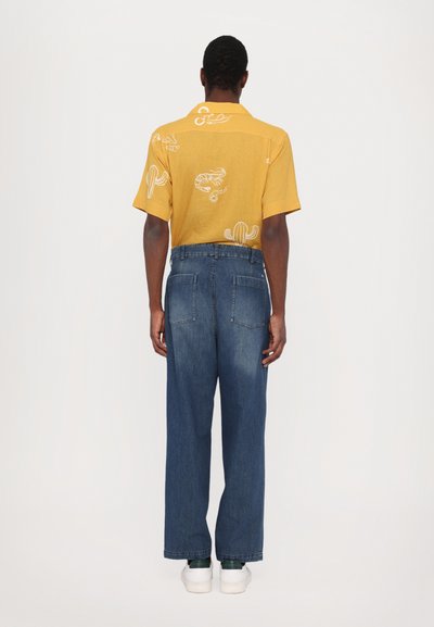 Gul t-skjorte med kort erme, prydet med hvite kaktus- og reke-mønstre, kombinert med vide blå jeans og hvite sneakers, sett fra ryggen.