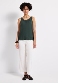 Top sin mangas de color verde oscuro con detalles fruncidos en el escote, combinado con pantalones blancos holgados y sandalias negras de mimbre.