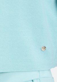 Lichtblauwe gebreide top met een soepele textuur en een zichtbaar metallic drukknopaccent nabij de zoom. Zachte en aansluitende pasvorm.