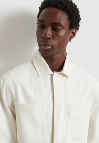 Giovane uomo con capelli corti e ricci, indossa una giacca color crema con due tasche frontali, guarda verso il basso e di lato.