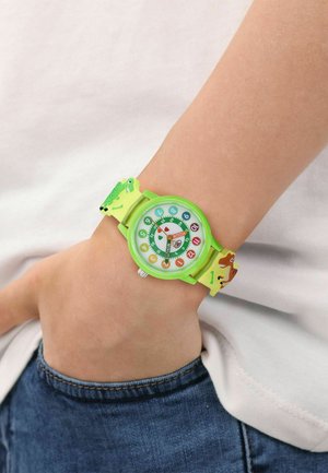 Child's green wristwatch with colorful numbers and animal designs on the strap, worn on the left wrist over a white shirt and blue jeans.