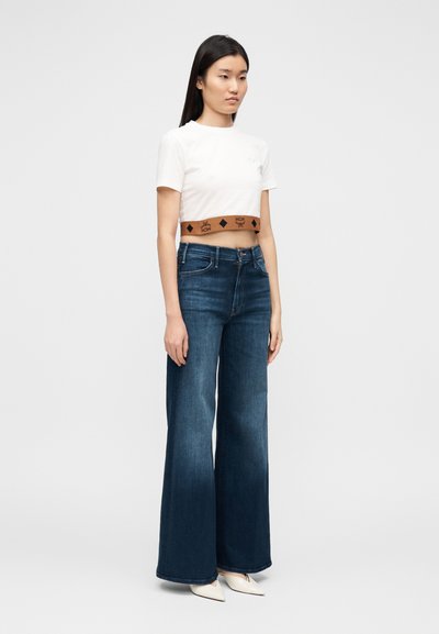 Κοντοκομμένο άσπρο μπλουζάκι με καφέ ζώνη με σχέδια, συνδυασμένο με σ κούρο μπλε παντελόνι καμπάνα. Το μοντέλο φοράει μυτερά άσπρα παπούτσια.