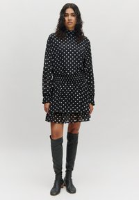 Schwarzes Kleid mit Punkten, tailliertem Oberteil, gerüschtem Saum und langen Ärmeln. Kombiniert mit knielangen schwarzen Stiefeln. Glatte Stofftextur.