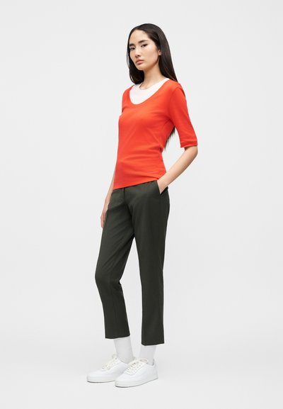 Κόκκινο τοπ με κοντά μανίκια από πάνω από ένα λευκό πουκάμισο, συνδυασμένο με σκούρο πράσινο παντελόνι και λευκά αθλητικά παπούτσια. Το μοντέλο στέκεται με τα χέρια στις τσέπες.