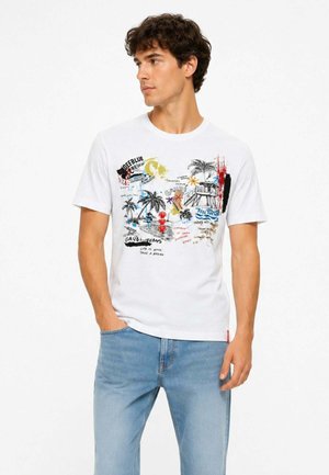Giovane uomo con capelli scuri e ricci che indossa una t-shirt bianca con illustrazioni colorate di spiaggia e palme e jeans azzurri.