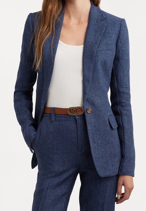 Femme portant un costume bleu marine cintré avec un blazer à un bouton, un débardeur blanc et une ceinture marron avec une boucle dorée, debout avec une main dans la poche.