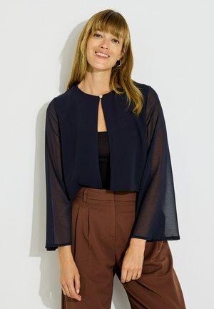 Femme souriante aux cheveux blonds et raides porte un chemisier court bleu marine transparent à manches longues et un pantalon plissé marron taille haute.