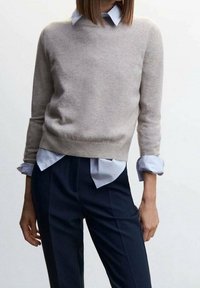 Béžový vlněný svetr s kulatým výstřihem a dlouhými rukávy, vrstvený přes světle modrou košilí s límečkem. Námořnické kalhoty s přesně vyžehlenými záhyby.