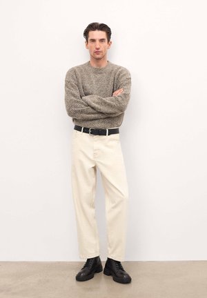 Pantaloni din bumbac bej, pulover texturat gri, curea neagră și bocanci negri cu șiret. Modelul stă cu brațele încrucișate pe un fundal simplu.