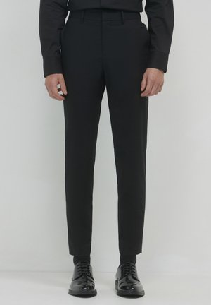 Schwarze maßgeschneiderte Hose mit schmalem Schnitt, aus glattem Stoff gefertigt, mit Gürtelschlaufen und kombiniert mit glänzenden schwarzen Kleiderschuhen.